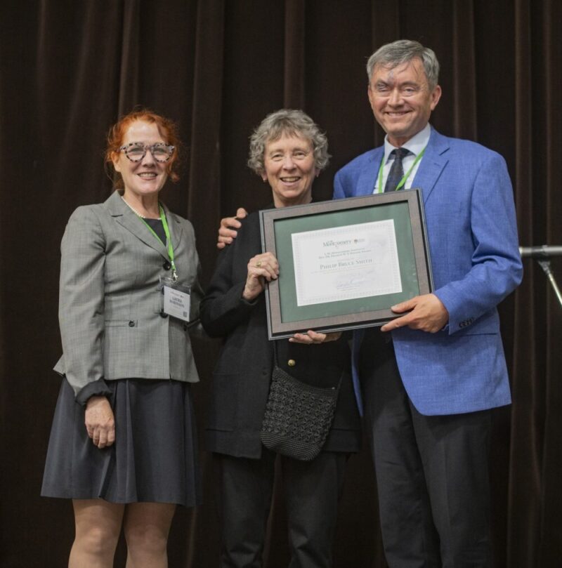 two women present a framed award to a man in a blue jacket