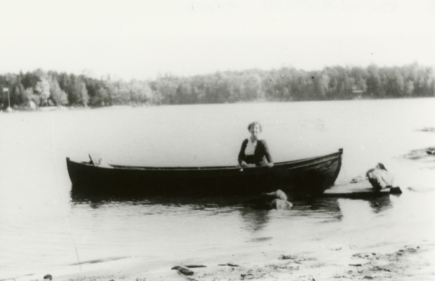 black and white image of a woman in dark clothing in a canoe on a small lake
